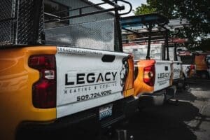 Three yellow service trucks with "Legacy Heating & Cooling" branding and contact info are parked in a row in a lot.