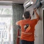 A technician in an orange "Legacy Heating & Cooling" shirt installs or repairs a wall-mounted air conditioning unit indoors, similar to work done on the University of Idaho HVAC project.