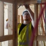 A construction worker in a yellow safety vest and hard hat installs wiring inside a wooden building frame for the University of Idaho HVAC project.